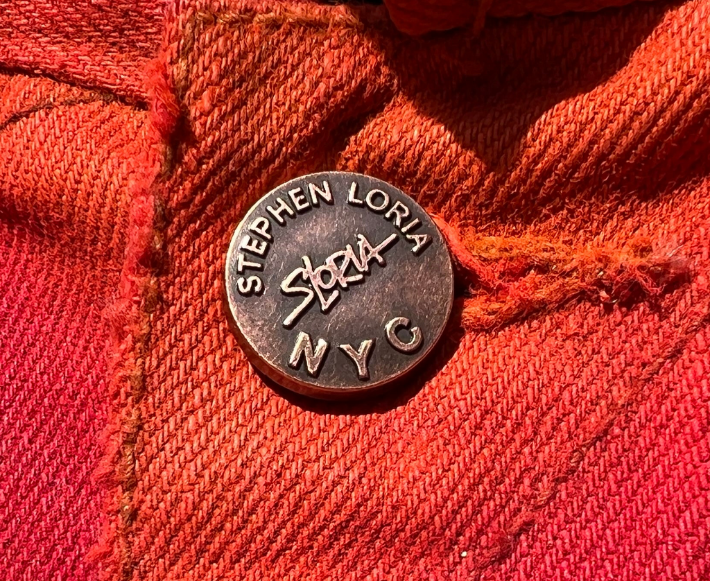 Round metal badge with 'Stephen Loria' branding on a red fabric background