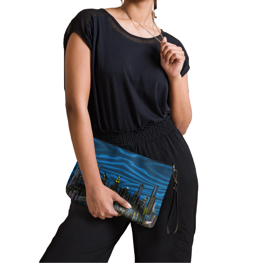 Woman holding a blue Loria NYC Art clutch with a cityscape design against a striped wall.