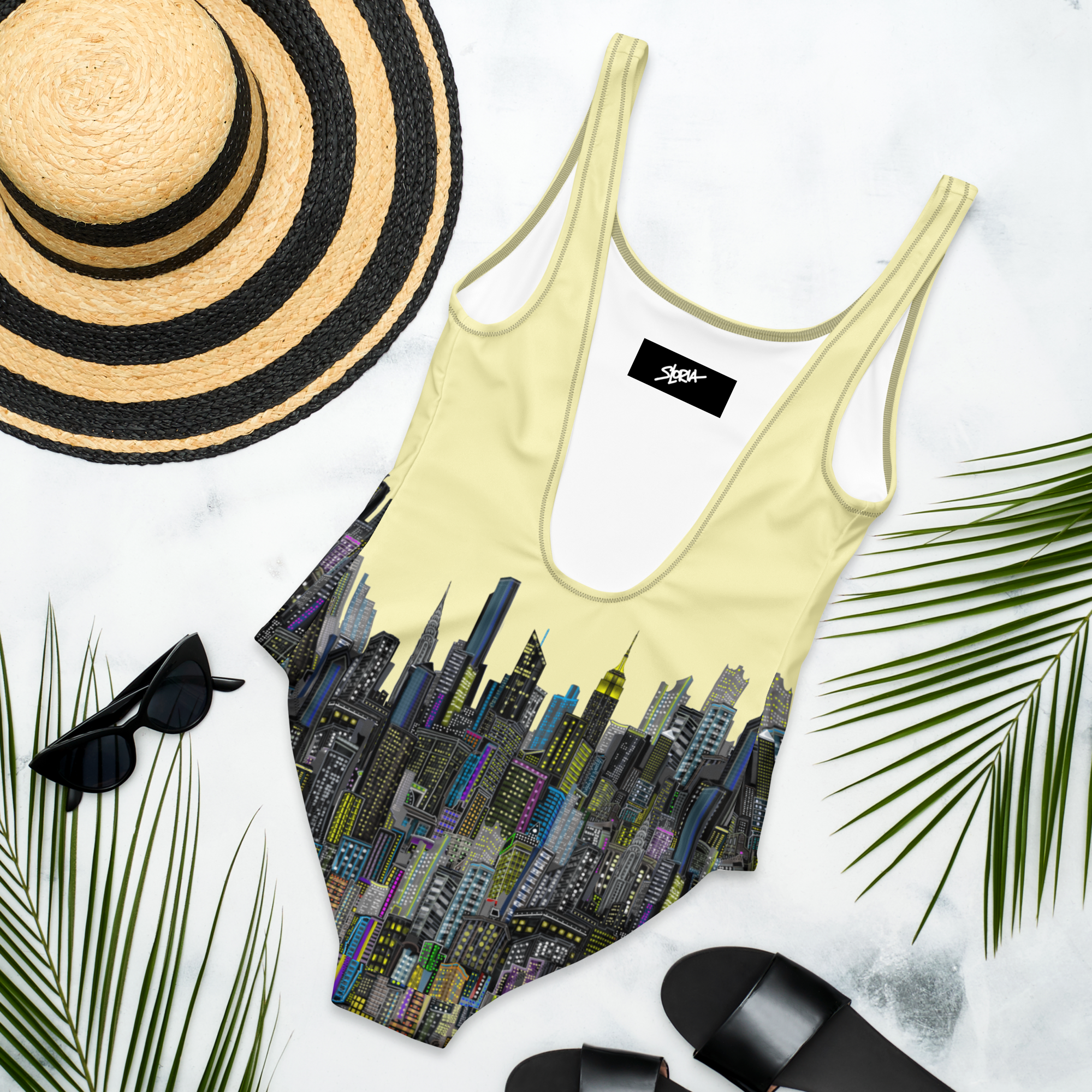Back of a Loria NYC Art Yellow swimsuit with cityscape design, black and straw hat, sunglasses, and black sandals on a white background.