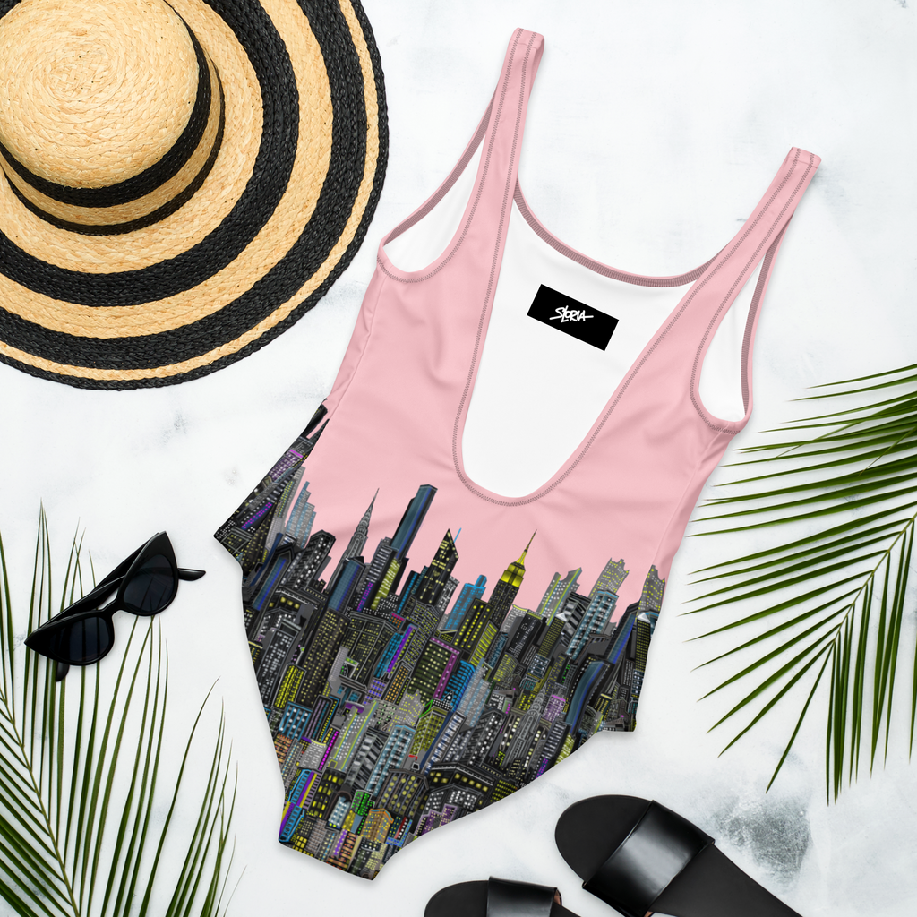 Back of a Pink Loria NYC Art swimsuit with cityscape design, black and straw hat, sunglasses, and black sandals on a white background.