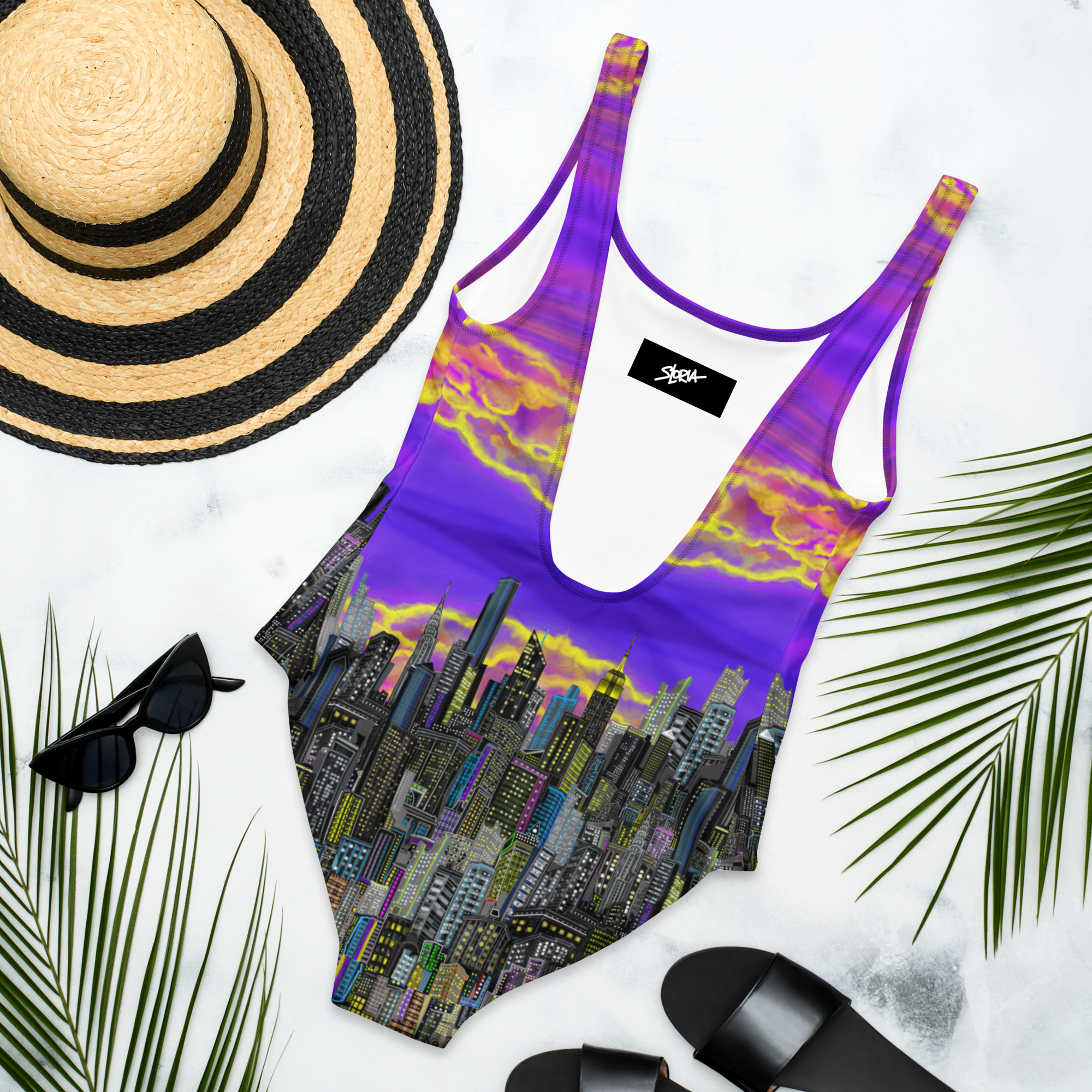 Purple swimsuit with cityscape Loria NYC Art design, sun hat, sunglasses, and sandals on a white background.