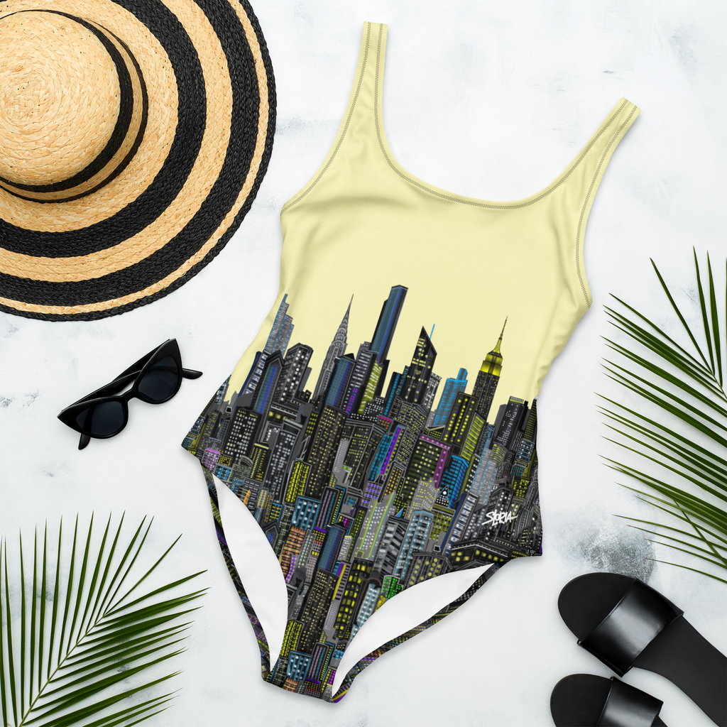 One-piece swimsuit with Loria NYC Art cityscape design, sun hat, sunglasses, and sandals on a light background