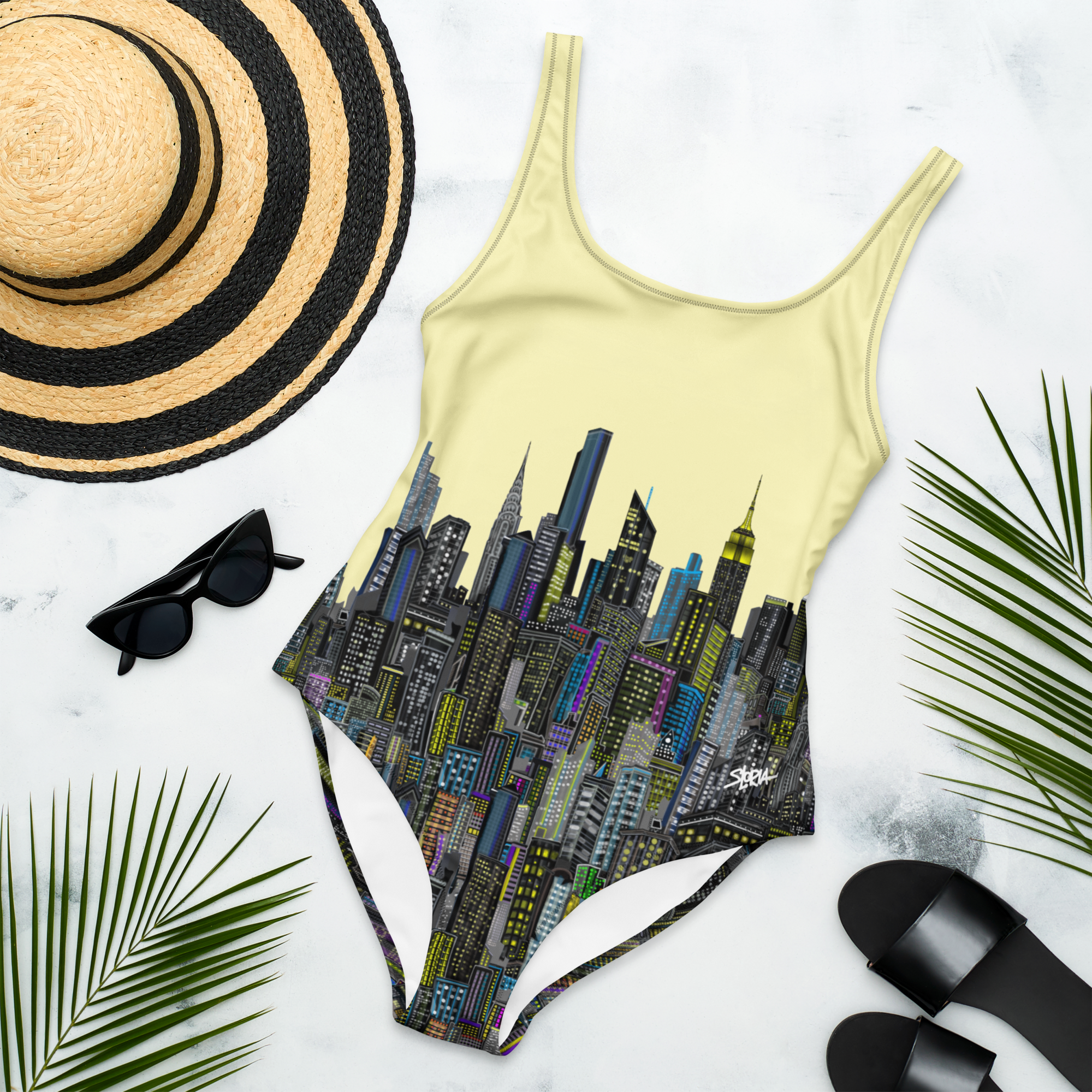 One-piece swimsuit with Loria NYC Art cityscape design, sun hat, sunglasses, and sandals on a light background
