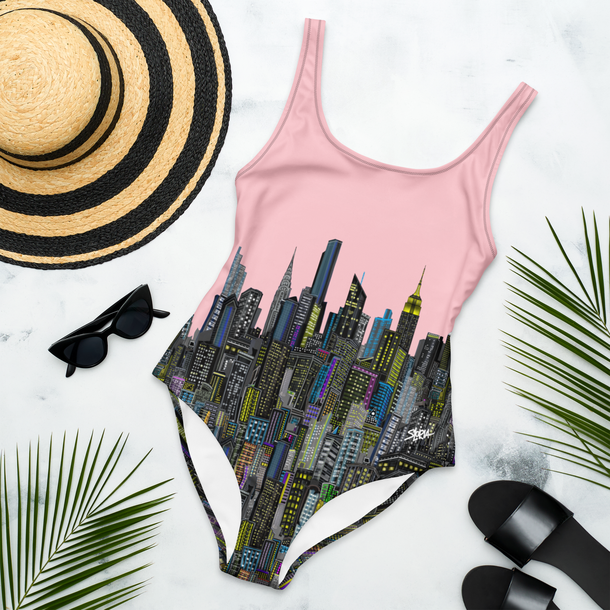 Pink cityscape Loria NYC Art swimsuit with a straw hat, sunglasses, and sandals on a white background