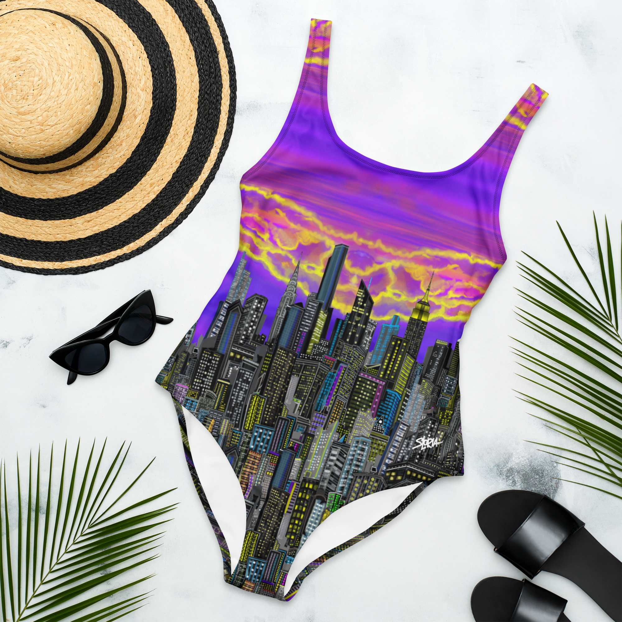 One-piece swimsuit with Loria NYC Art cityscape design, sun hat, sunglasses, and sandals on a white background