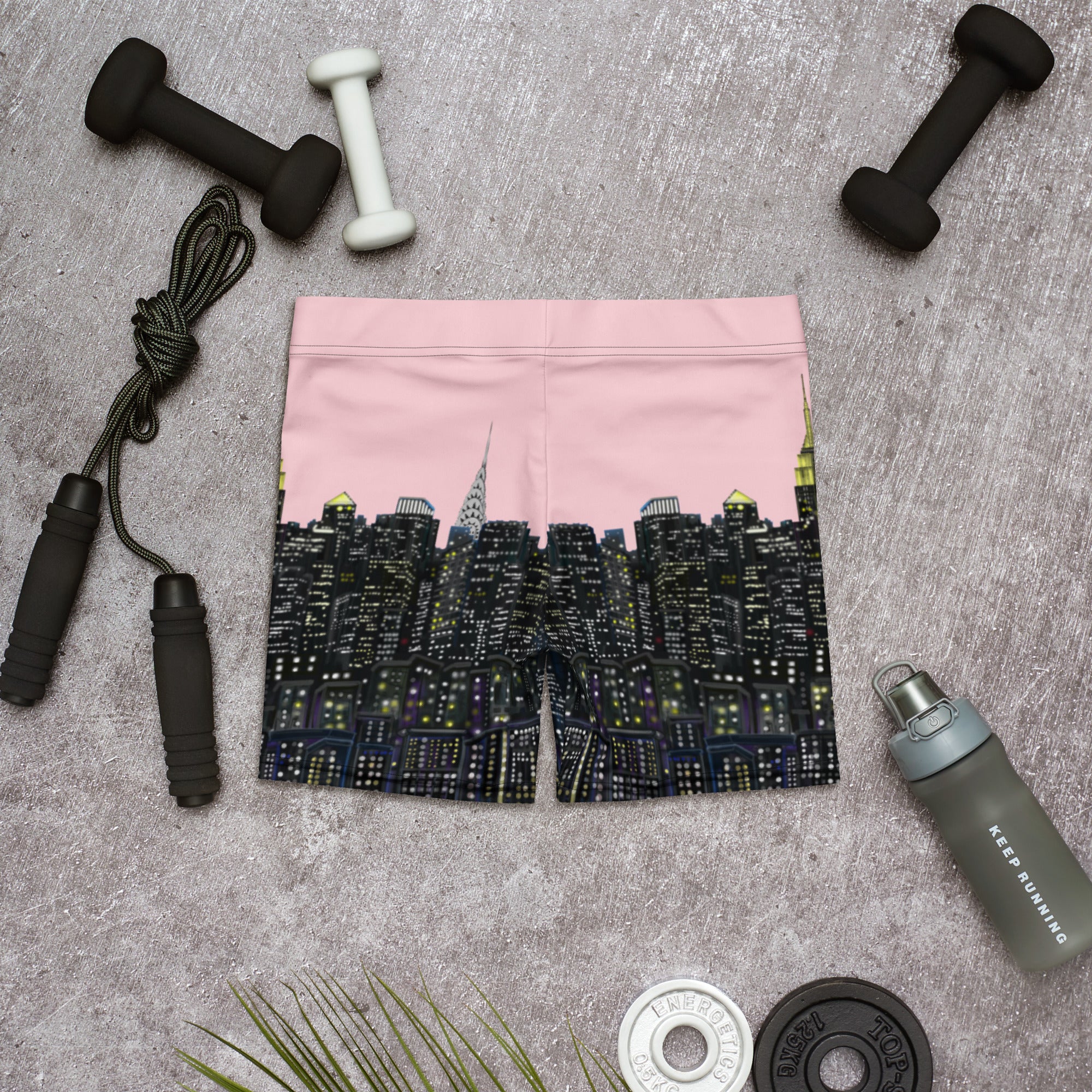 Pink athletic shorts with  Loria NYC Art skyline design on a gray surface with fitness equipment.