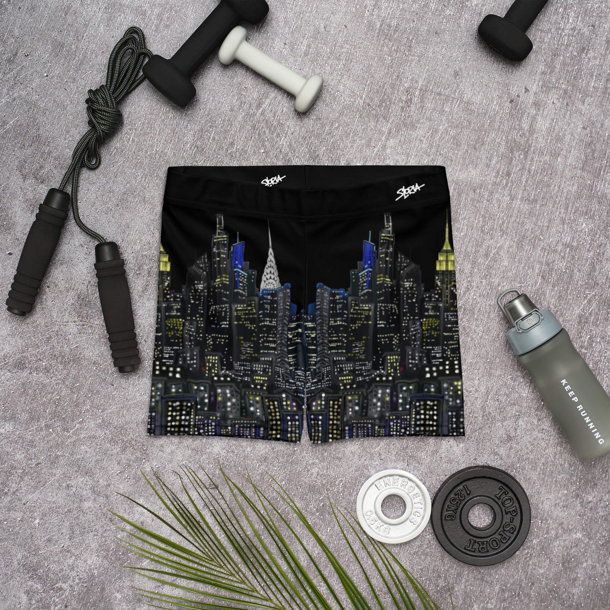 Black swim shorts with cityscape Loria NYC Art design on a gray surface with fitness equipment.