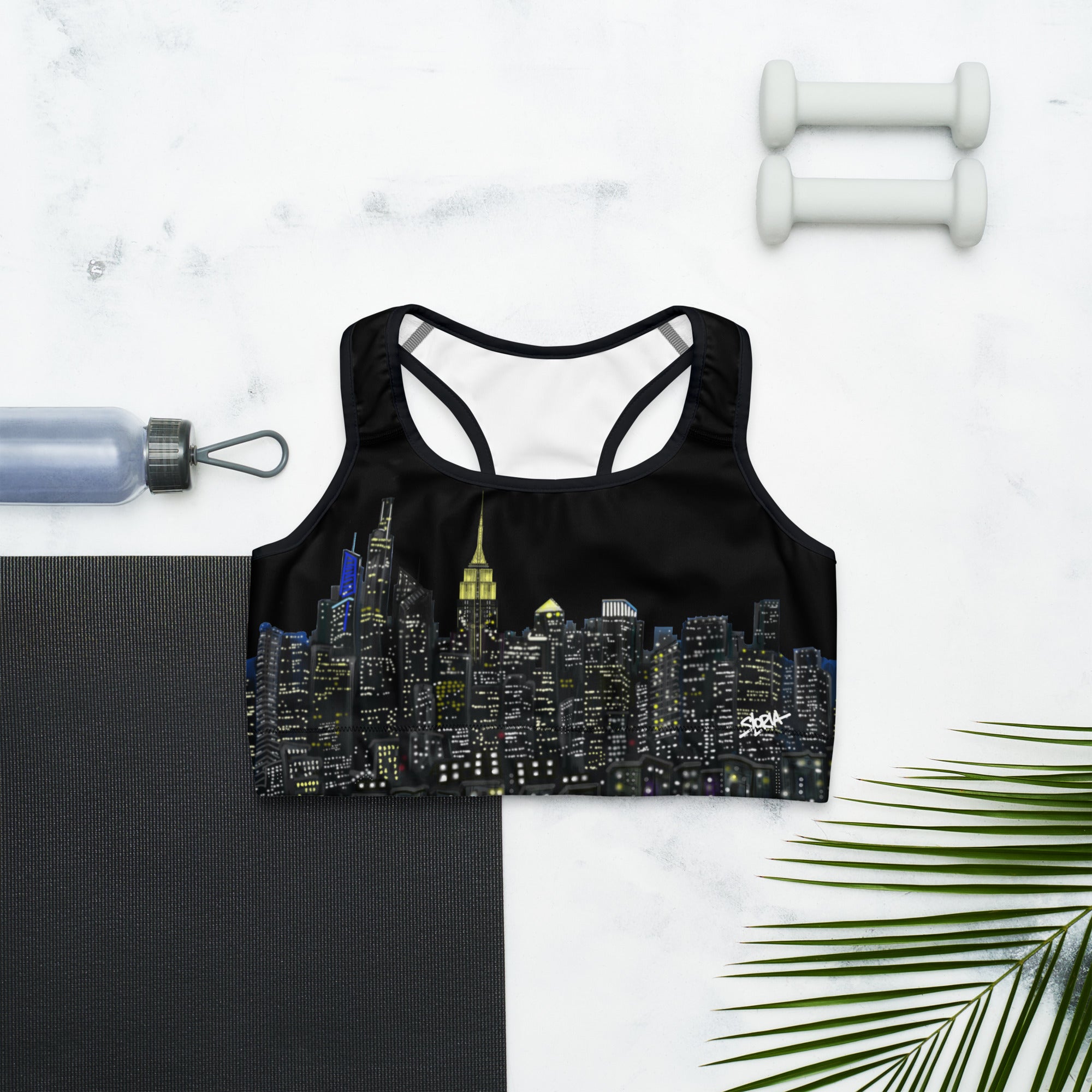 Black sports bra with cityscape design on a marble surface with a yoga mat, water bottle, and dumbbells.