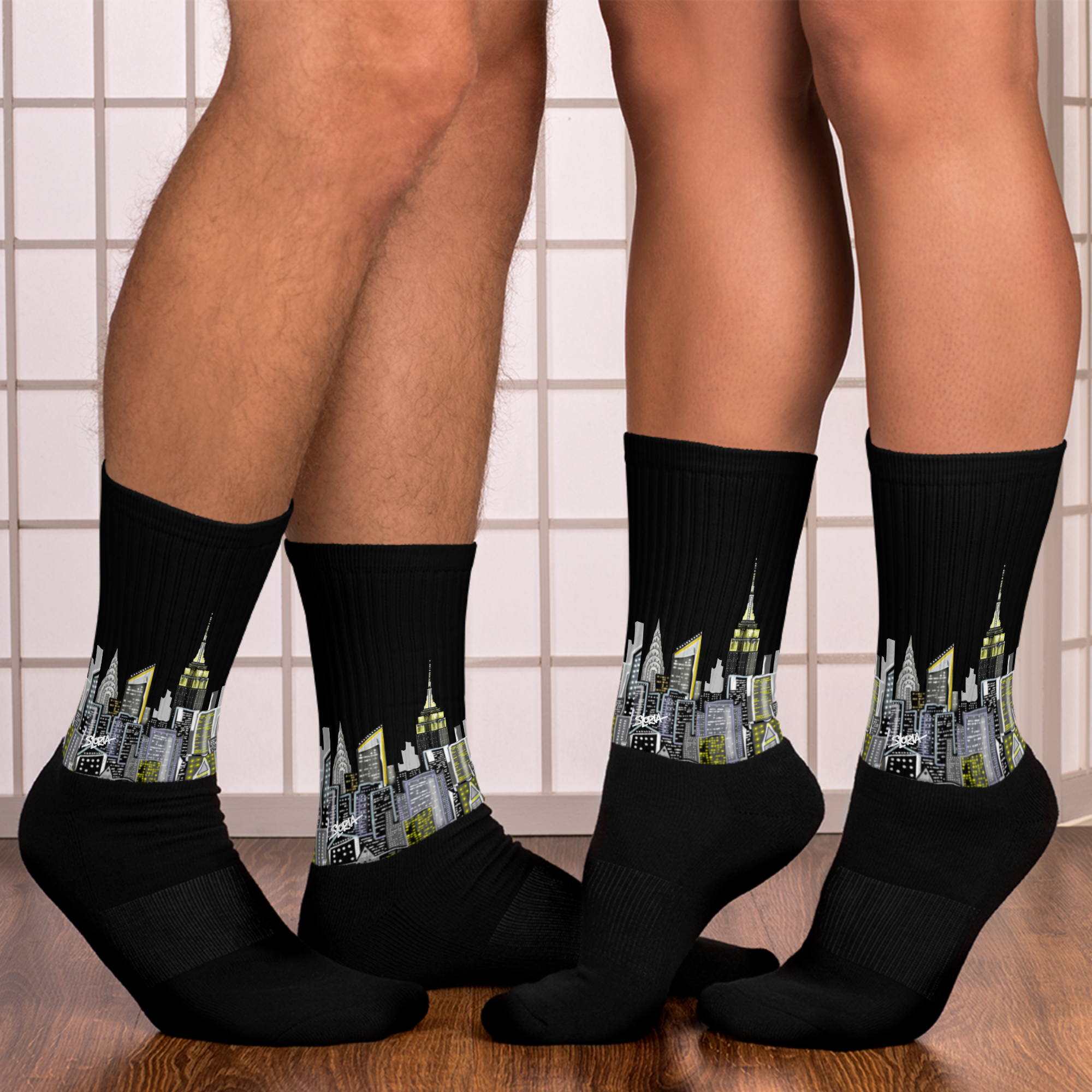 Black Loria NYC Art socks with cityscape design worn by two people on a tiled floor.
