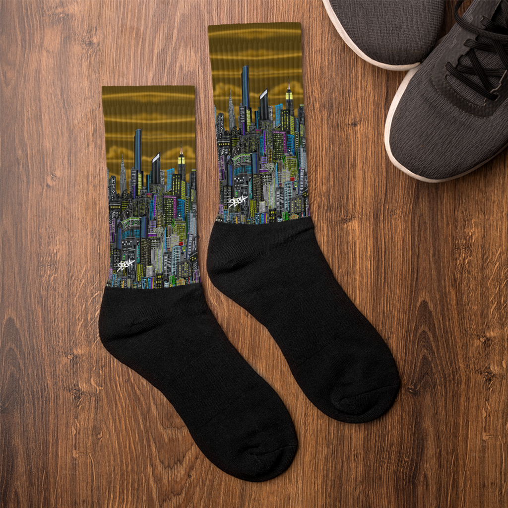 Black socks with Loria NYC Art cityscape design on a wooden surface next to a pair of shoes.