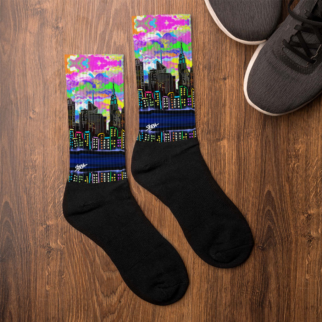 Black socks with colorful cityscape Loria NYC Art design on a wooden surface next to a pair of shoes.
