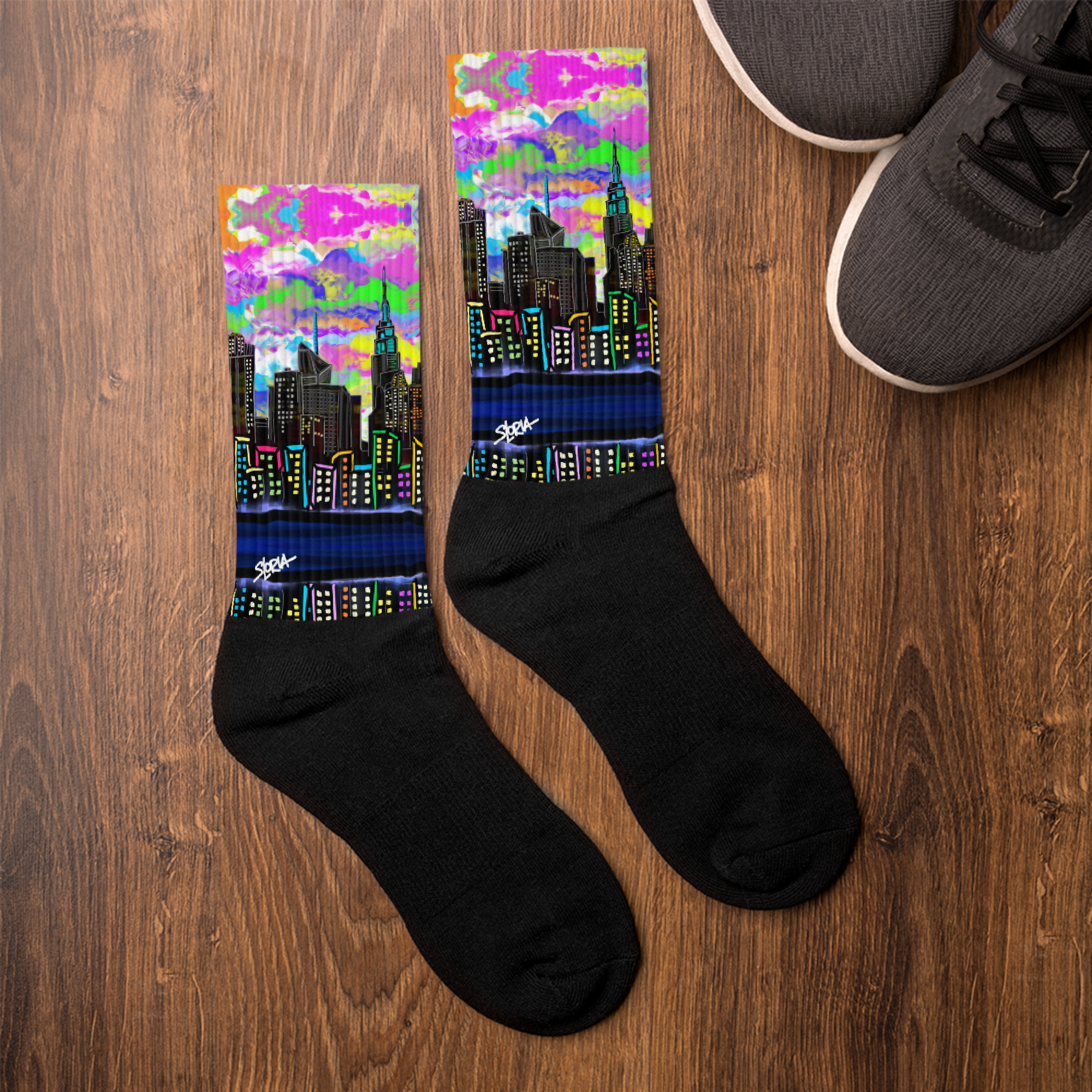 Black socks with colorful cityscape Loria NYC Art design on a wooden surface next to a pair of shoes.