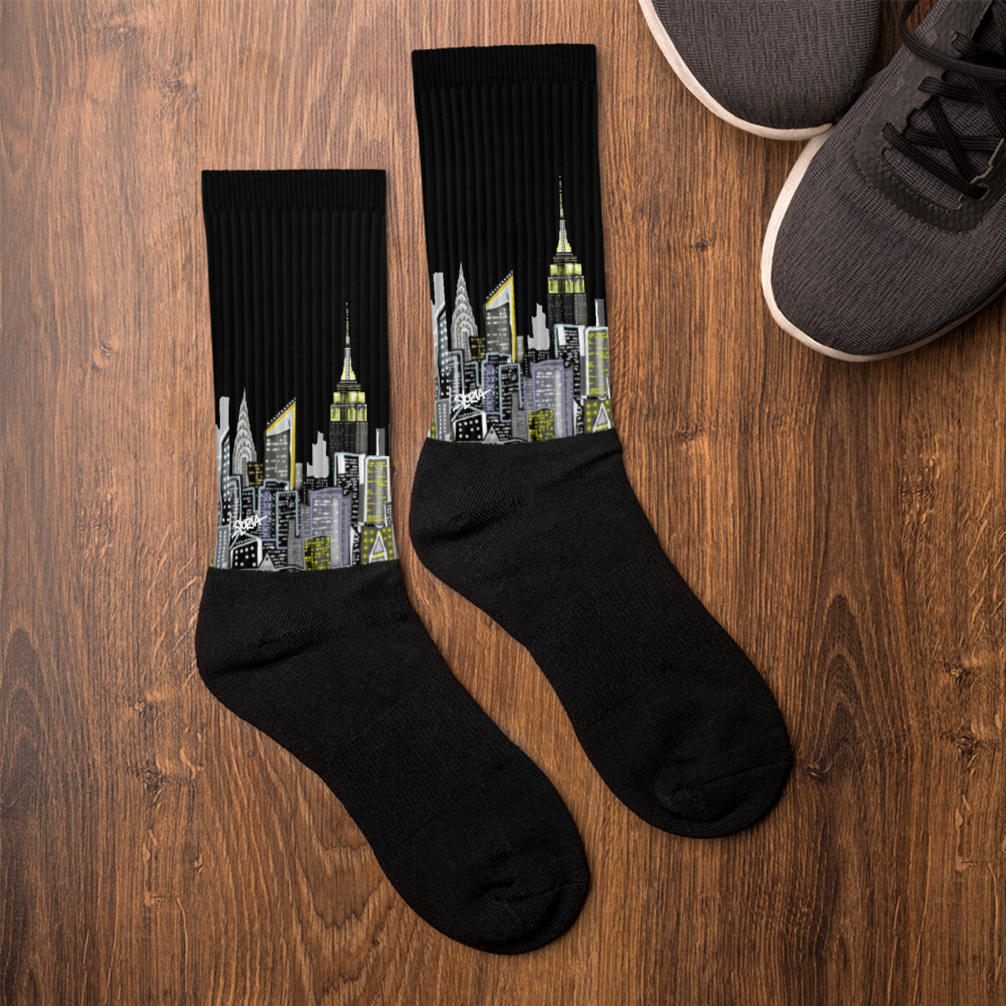 Black socks with cityscape Loria NYC Art design on a wooden surface next to a pair of shoes.