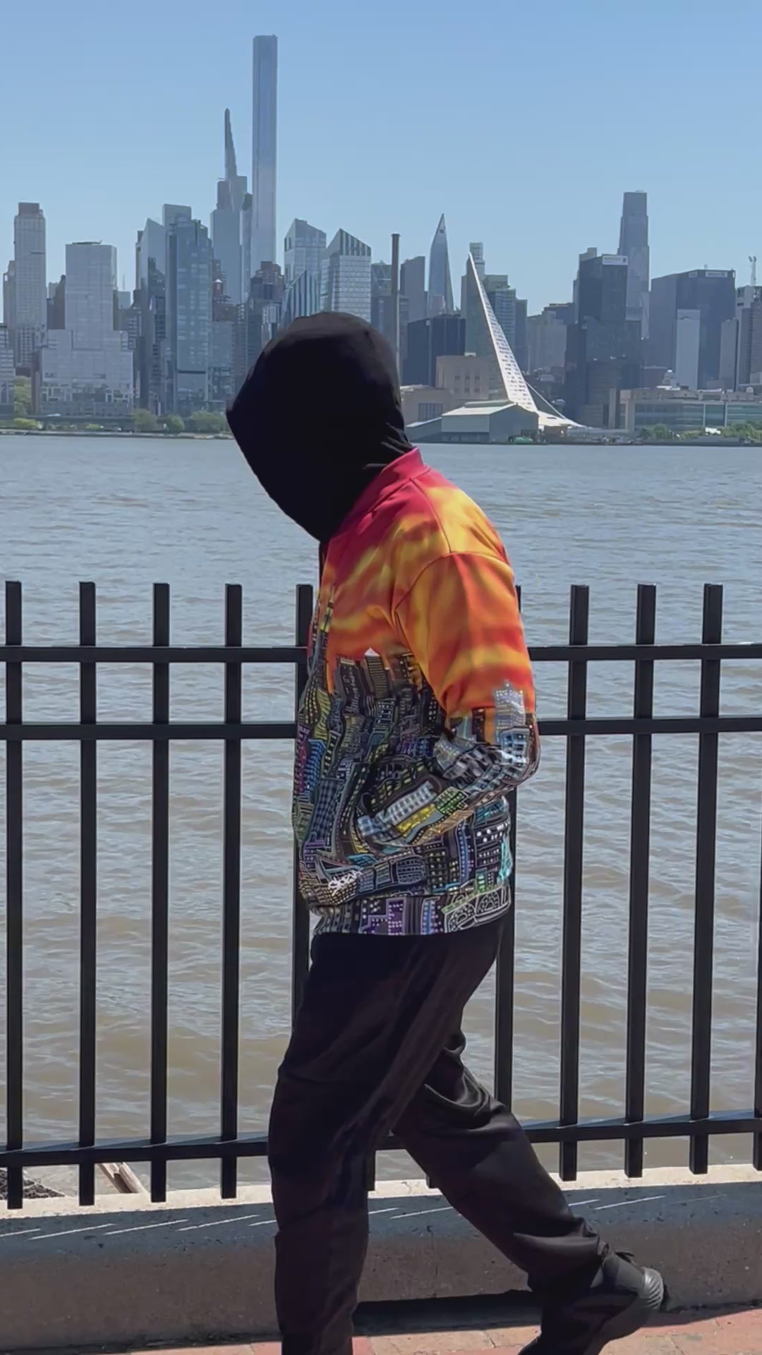 A person walking along a view of the skyline wearing a vibrant Loria NYC Art jacket with a detailed city skyline graphic across the back, featuring a fiery orange and yellow gradient sky. 