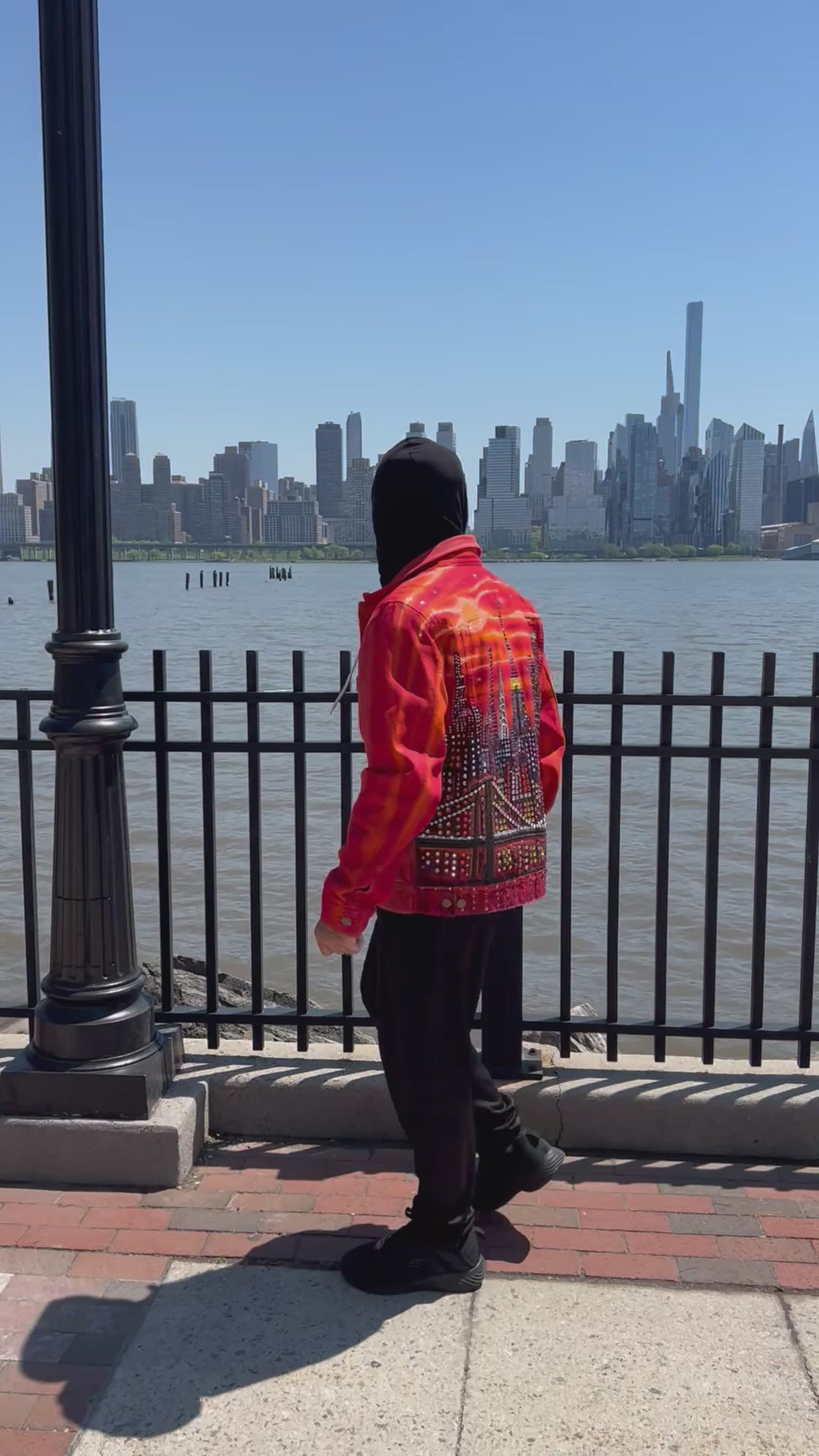 Person in a video wearing an orange Loria NYC Art jacket looking at  the city skyline in the background.