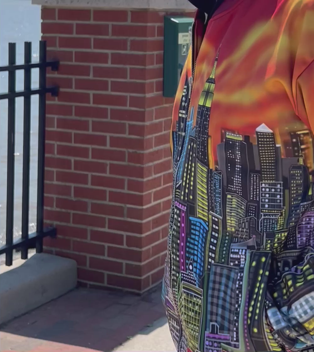 A video of a person viewed from behind wearing a vibrant Loria NYC Art jacket with a detailed city skyline graphic across the back, featuring a fiery orange and yellow gradient sky. The person is standing outdoors near a railing.