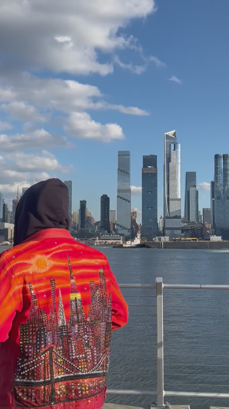 Video of a person wearing a vibrant orange Loria NYC Art jacket looking at  the skyline in the background.