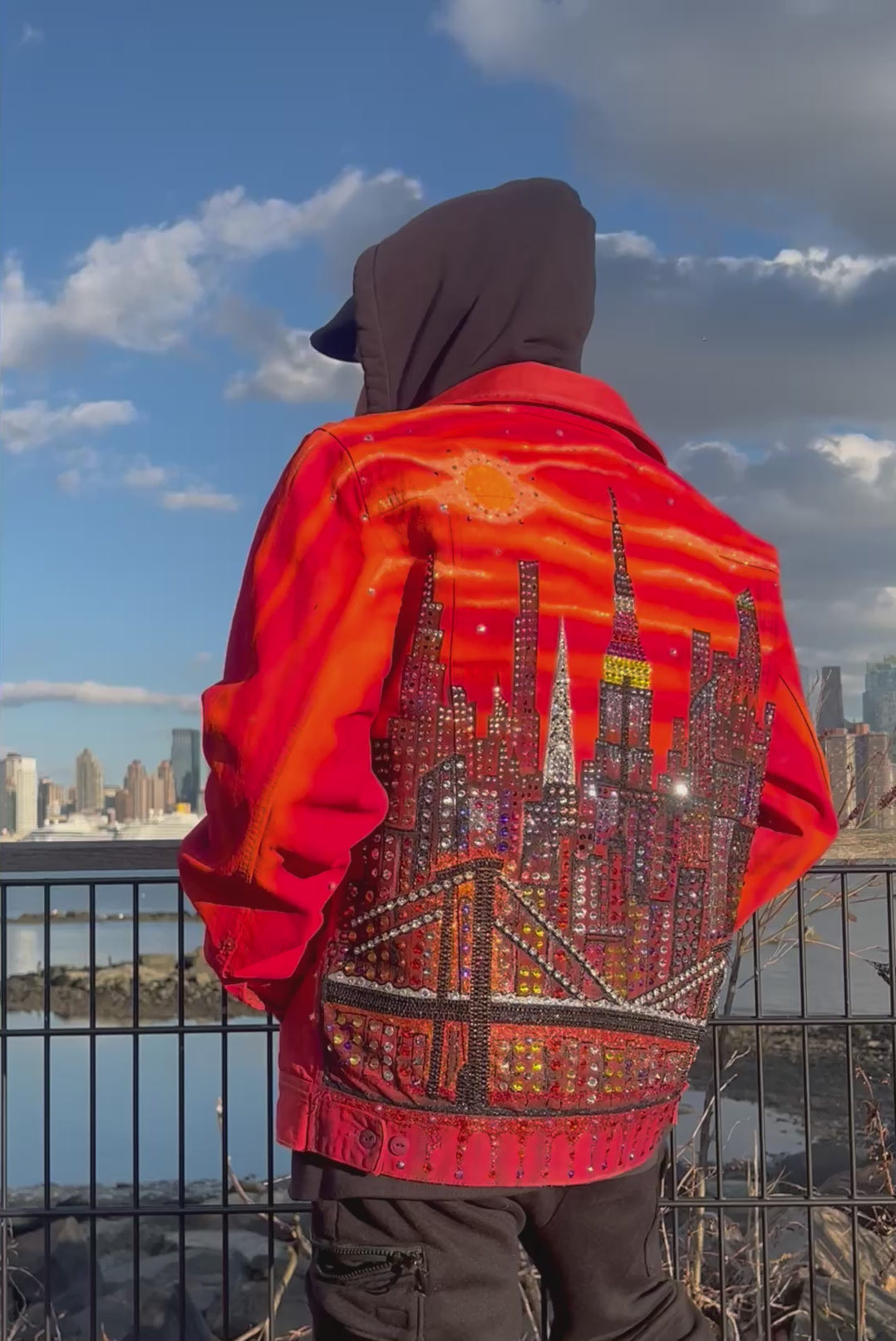 Man posing in a video wearing a vibrant orange Loria NYC Art jacket looking at  the city skyline in the background.