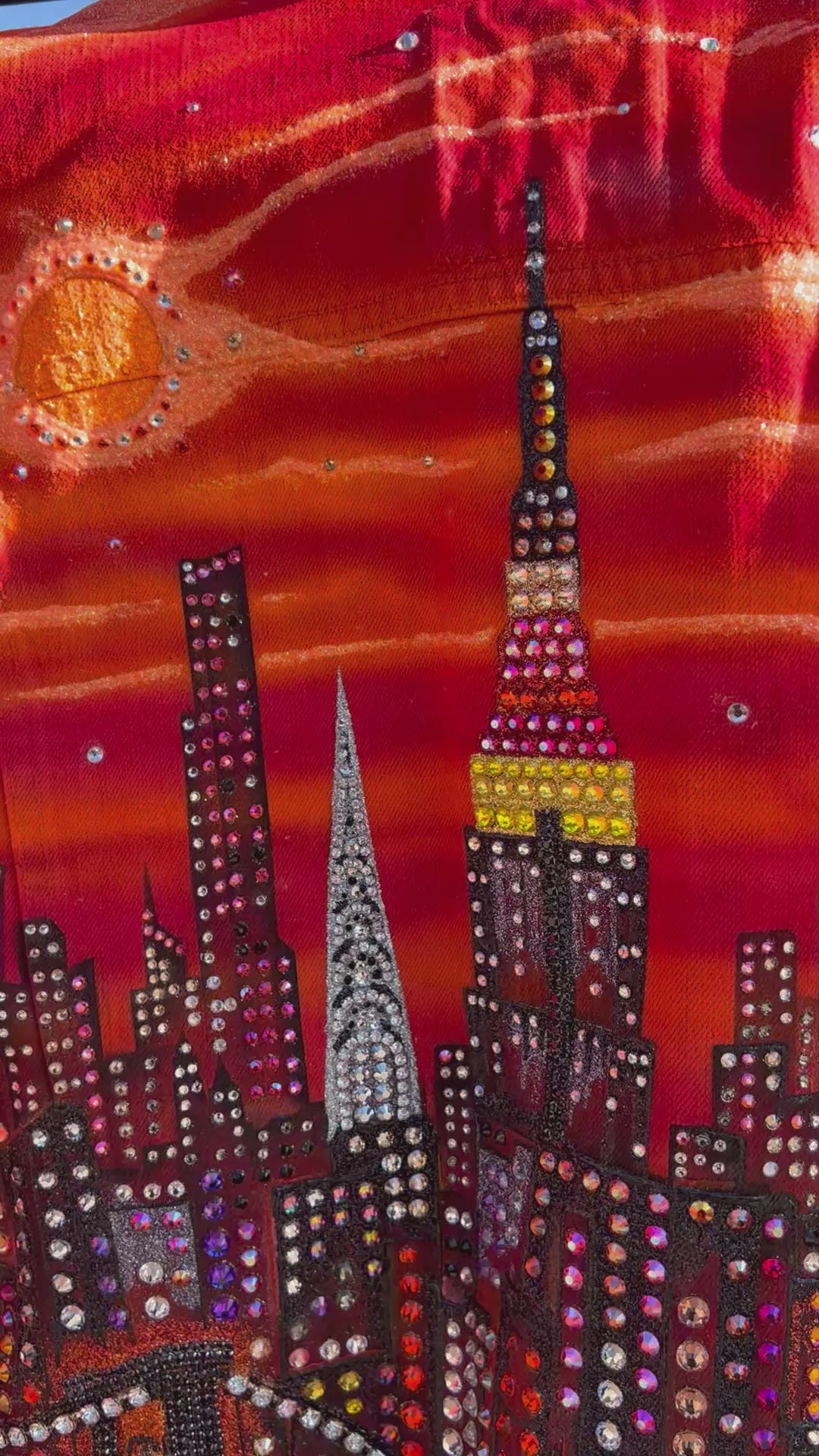 Video of an orange Loria NYC Art jacket in front of the city skyline along a railing.