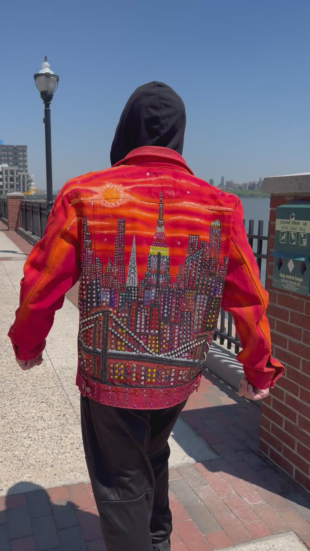 Person walking wearing a vibrant orange Loria NYC Art jacket looking at  the city skyline in the background.