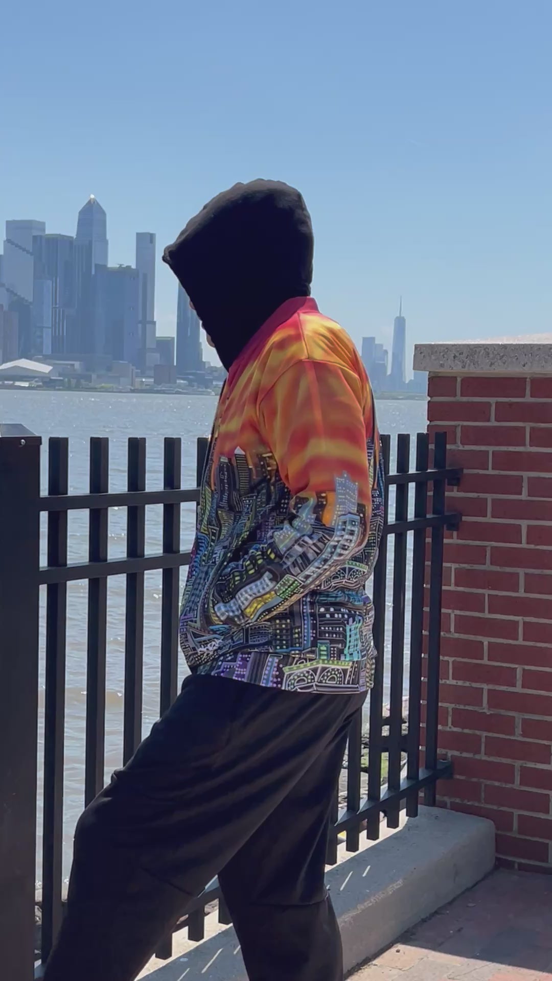 A person in a video viewed from behind wearing a vibrant Loria NYC Art jacket with a detailed city skyline graphic across the back, featuring a fiery orange and yellow gradient sky. The person is standing outdoors near a railing with a skyline view.