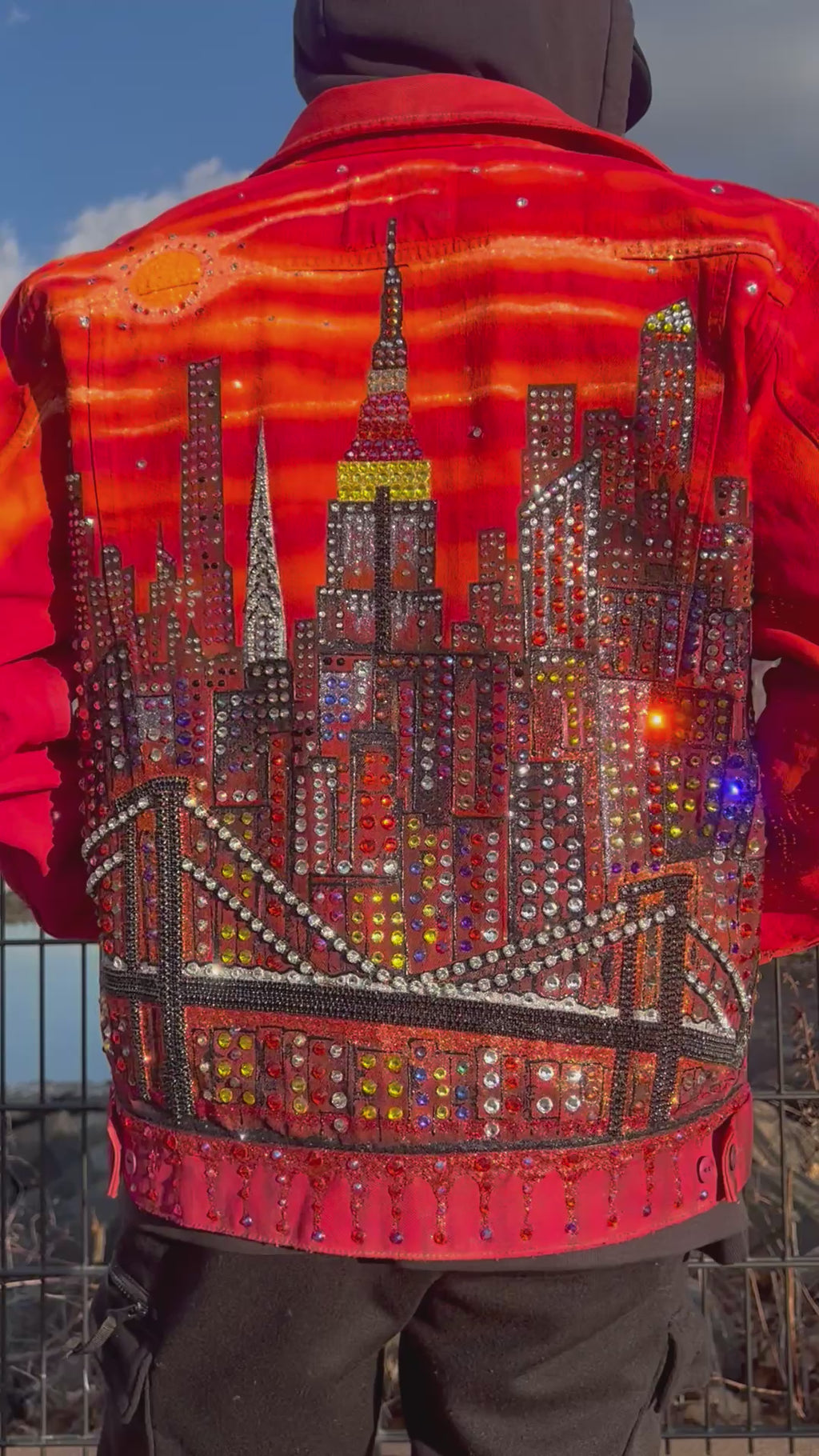 Man in a video wearing a vibrant orange Loria NYC Art jacket with the city skyline in the background.