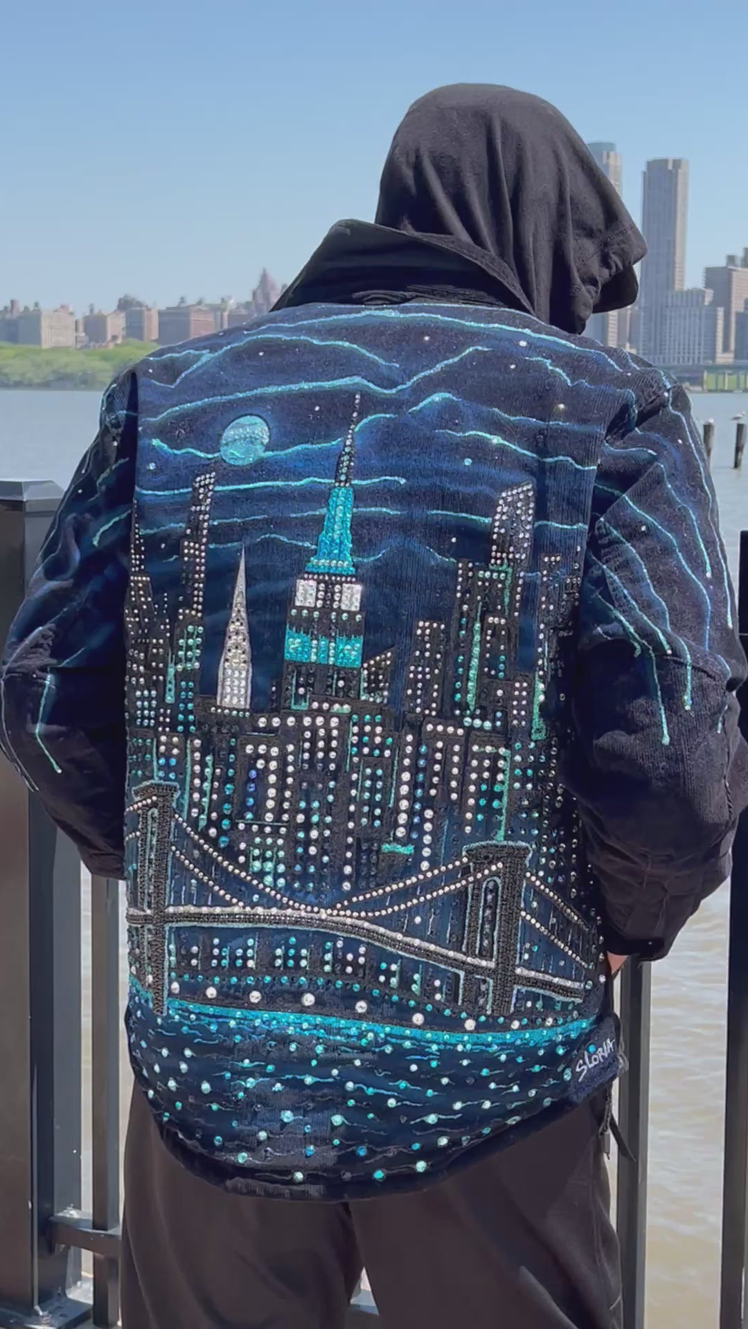 Man wearing a blue jacket with Loria NYC Art on back in a video with skyline in the background.