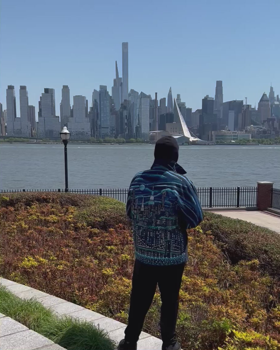 Person walking with a Blue colorful jacket with Loria NYC Art on the back with the city skyline in the background.