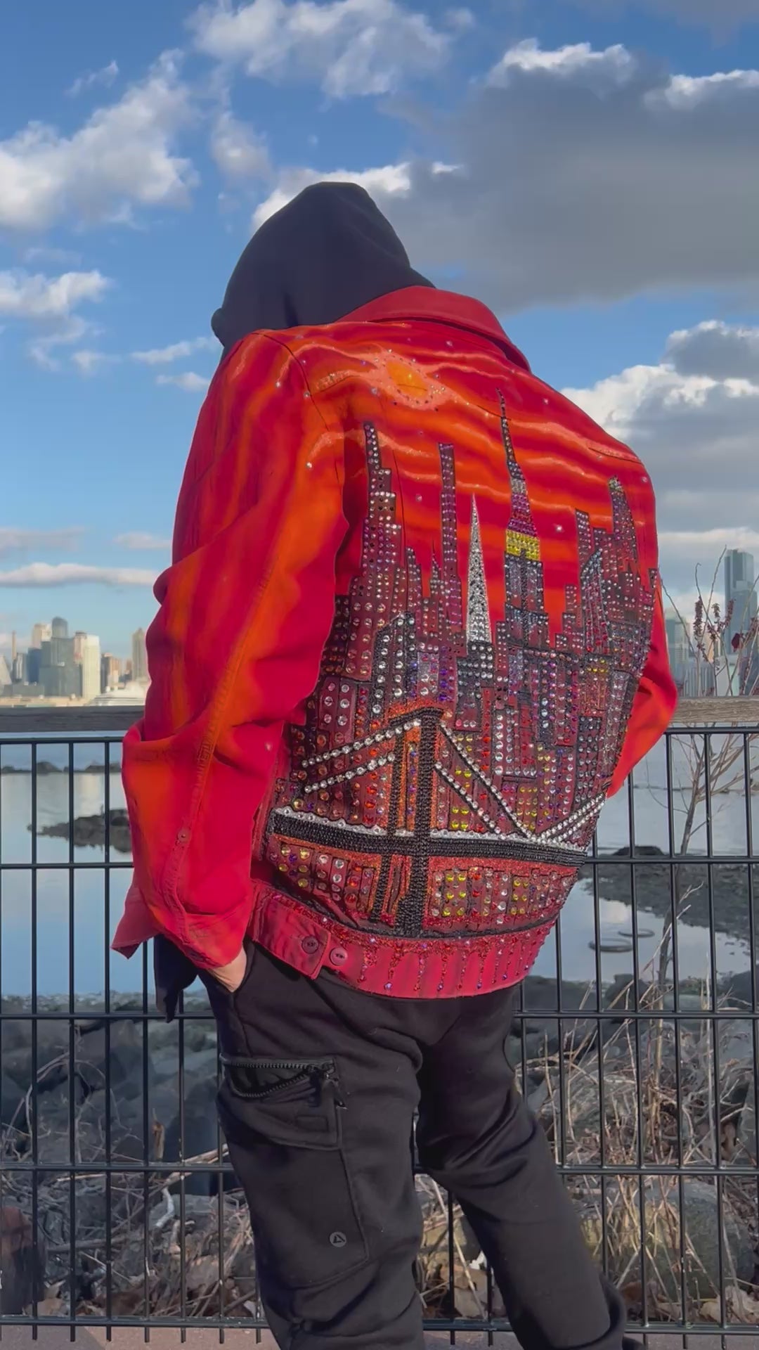 Person wearing an orange Loria NYC Art jacket looking at  the city skyline in the background.