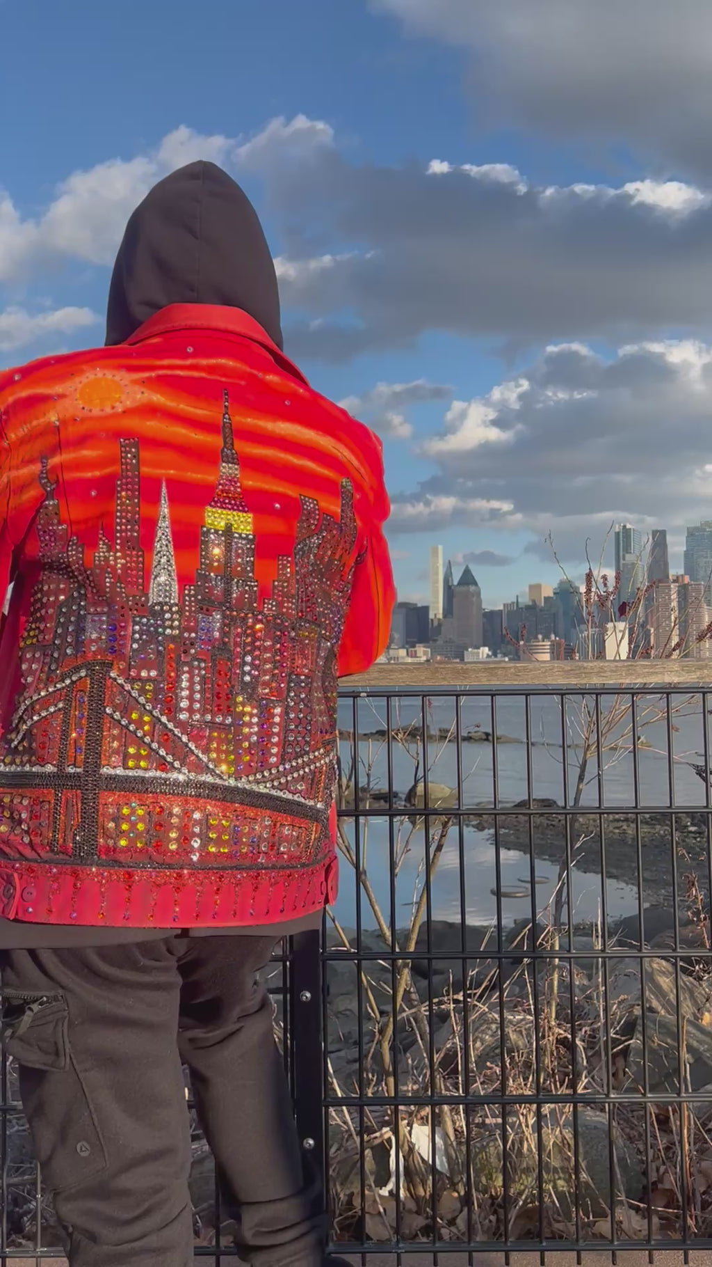 Man standing in a video wearing a vibrant orange Loria NYC Art jacket looking at  the city skyline in the background.